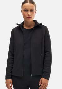 Chaqueta negra con cremallera y un cuello suave, hecha de material polar, cuenta con dos bolsillos laterales y una textura suave, llevada sobre una camiseta negra.