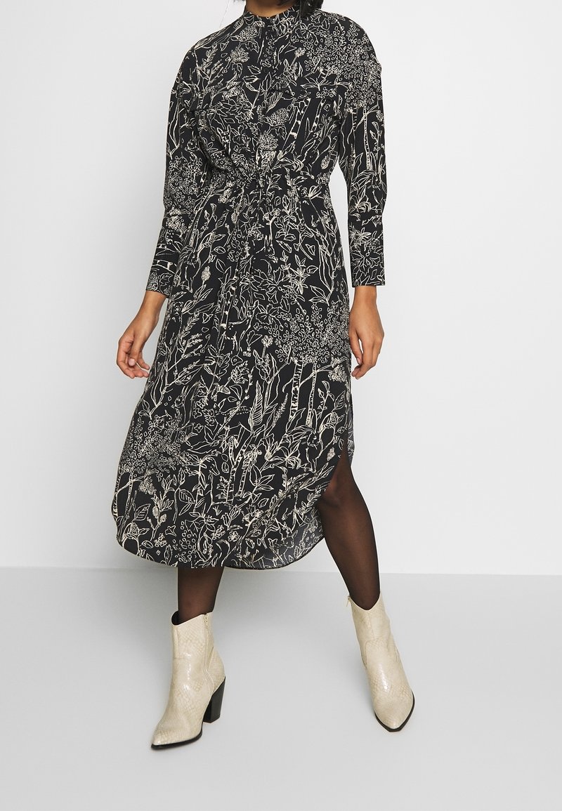 Abito midi nero con stampa floreale bianca, maniche lunghe, colletto alto, vita cintrata, spacco laterale, abbinato a stivaletti beige texturizzati.