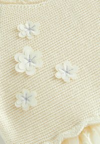 Tissu tricoté crème clair présentant trois fleurs décoratives en tissu avec des broderies argentées. Texture douce avec un bord festonné.
