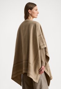 Poncho beige realizzato in un tessuto morbido con una forma rettangolare, caratterizzato da motivi testurizzati e un design drappeggiato. Maniche lunghe con polsini a costine.