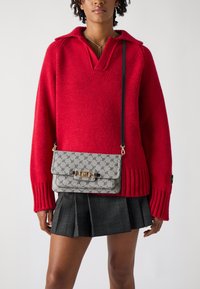 Gray patterned crossbody bag with a gold clasp, worn over a red oversized sweater and a black plaid skirt; textured knit detail visible.