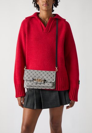 Grå mønstret crossbody taske med en guldkrog, båret over en rød oversized sweater og en sort plaid nederdel; tekstureret strikdetalje synlig.