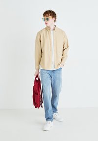 Camicia beige con bottoni, t-shirt bianca, jeans azzurri chiari, scarpe bianche, in mano uno zaino rosso. Capelli ricci, indossa occhiali da sole azzurri chiari.
