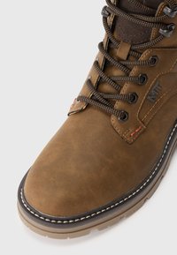 Bruine leren wandelschoen met zwarte en oranje veters, metalen oogjes, gestikte zool en gevoerde enkelkraag op een witte achtergrond.