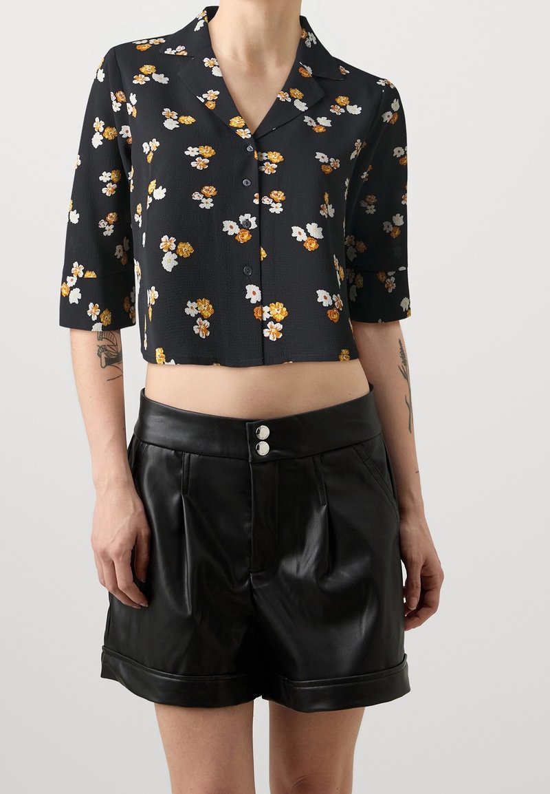 Camicetta nera a fiori con maniche corte, caratterizzata da fiori gialli e bianchi. Abbinata a pantaloni shorts alti in pelle nera con bottoni argentati.