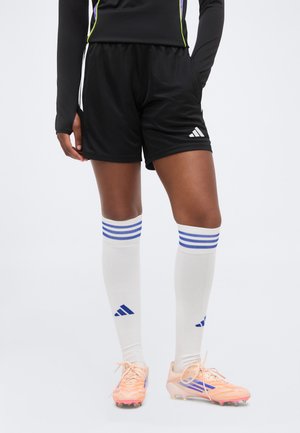 Persona con pantalones cortos negros Adidas, calcetines blancos hasta la rodilla con rayas azules y botas de fútbol de color durazno, de pie sobre un fondo liso.