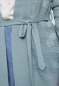 Ljusblå stickad cardigan med öppen front, strukturerat mönster och midjeband. Har mjukt, andningsbart tyg och intrikat sömnad.