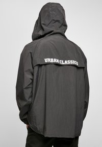Black hooded jacket with a textured finish, featuring "URBAN CLASSICS" printed in white on the back. Loose fit with elastic cuffs.