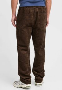 Braune Cordhosen mit lockerer Passform, vertikalen Rippungen, einem elastischen Bund und einer Gesäßtasche. Getragen mit grauen Turnschuhen.