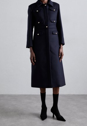Long manteau bleu marine foncé avec boutons dorés, poches poitrine et taille devant, porté avec des bottines chaussettes noires à talons hauts sur fond de sol gris.