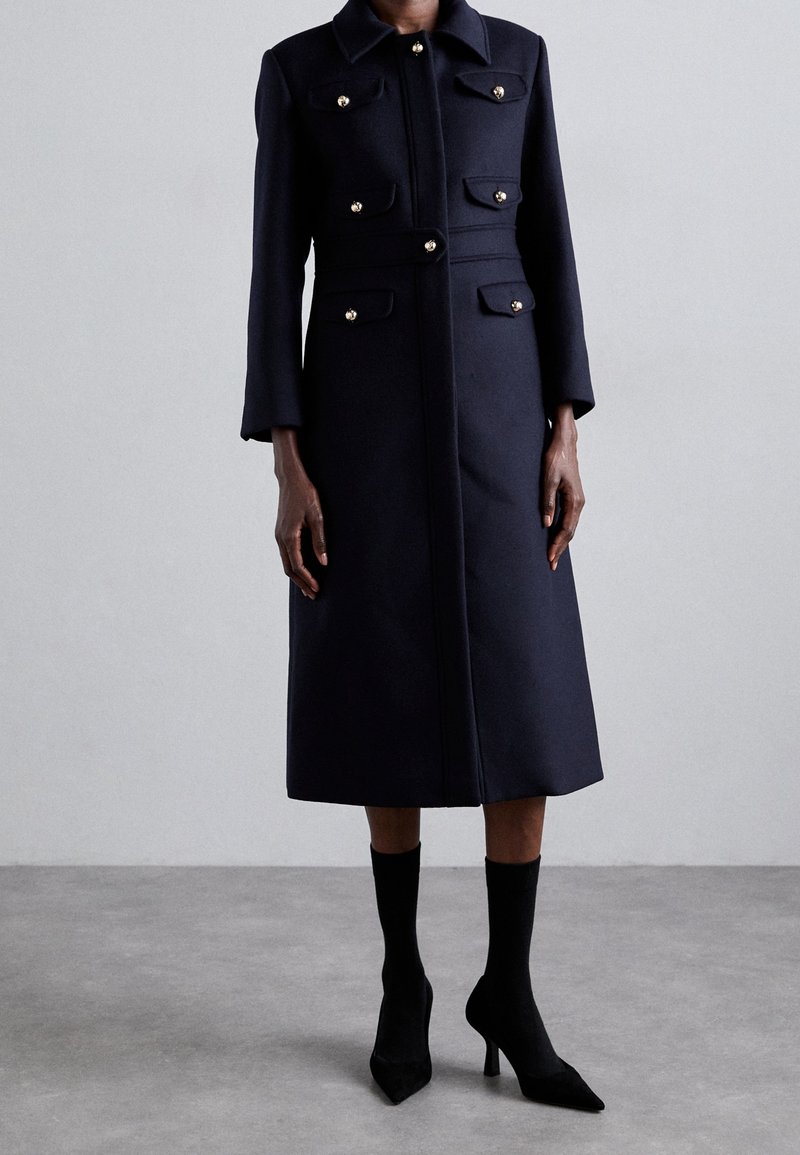 Long manteau bleu marine foncé avec boutons dorés, poches poitrine et taille devant, porté avec des bottines chaussettes noires à talons hauts sur fond de sol gris.