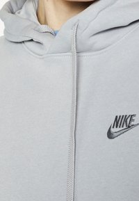 Nike šedá mikina s kapucí na šňůrku, měkkou texturou a černě vyšitým logem na hrudi.