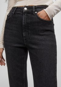 Jean en denim noir taille haute avec une texture lisse, poche avant et détail de bouton argenté. Coupe ajustée le long des jambes, sans usure visible.