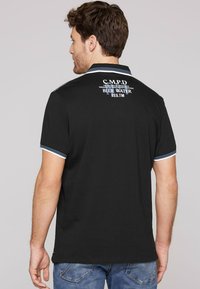 Czarny polo z kontrastującym niebiesko-białym kołnierzykiem, z nadrukowanym logo na plecach. Krótkie rękawy z akcentami w paski.