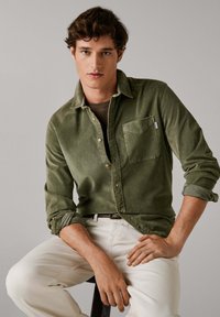 Camisa de pana verde oliva con cierre de botones, bolsillo en el pecho y mangas remangadas, combinada con pantalones en un tono crema claro. Tejido texturizado, diseño casual.