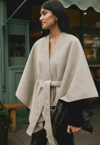 Beež villane kätikuub, millel on vöö, laiad varrukad ja V-kaelus. Kombineeritud musta nahk-käekotiga ja kantud ettepandud mustade pükstega.