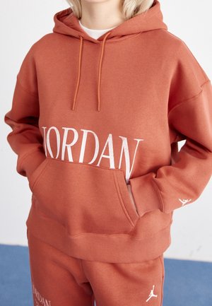 Personne portant un sweat à capuche Jordan couleur rouille et un pantalon assorti, les mains dans la poche du sweat, sur un fond uni.