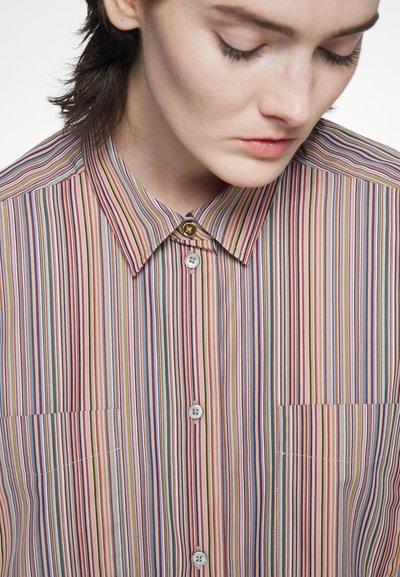 Camisa com riscas em tons pastel, apresentando um colarinho pontiagudo, dois bolsos frontais e um detalhe de botão dourado acima do botão superior.