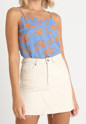 Kvinde iført en blå spaghetti-strap top med orange blomsterprint og en beige højtaljet denim nederdel med frynset kant.