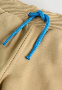Beige kokvilnas šorti ar ribotu jostu un zilu pītu mežģīti ar plastmasas galiem. Gluda tekstūra ar ikdienišķu dizainu.
