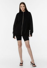 Czarna bluza z kapturem na zamek w mieszance bawełny, z przednią kieszenią i kapturem; w połączeniu z czarnymi szortami motocyklowymi i sandałami na obcasie.