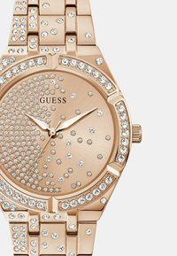Rožnato zlati uro s okroglo številčnico, okrašeno z rhinestones. Poliran kovinski pas, minimalistična številčnica in srebrne kazalke. Prikazana blagovna znamka 'GUESS'.