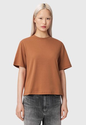 Femme aux longs cheveux blonds portant un t-shirt marron à manches courtes et un jean noir délavé, debout devant un fond gris uni.