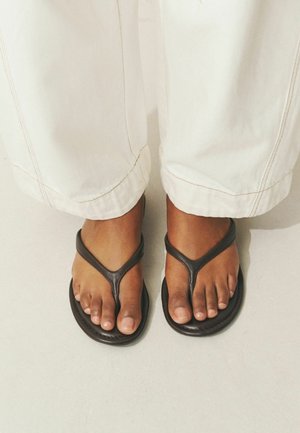 Pieds à la peau brune portant des sandales tongs marron foncé et un pantalon blanc à jambes larges, debout sur un sol clair.