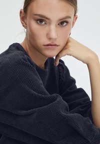 Dunkelblauer gerippter Strickpullover mit strukturierter Oberfläche und voluminösen Ärmeln. Das Modell stützt ihr Kinn auf die Hand.