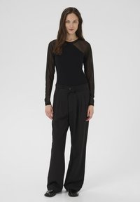 Mujer de pie con una blusa negra de manga larga transparente, pantalones negros holgados con cinturón y zapatos negros frente a un fondo claro liso.