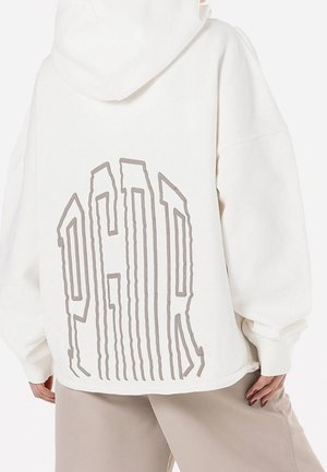 Person trägt einen weißen Hoodie mit Oversized-Passform und großem, stilisiertem Schriftzug "PGDR" auf dem Rücken, kombiniert mit beigen Hosen.
