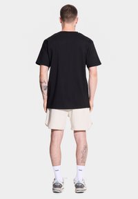 Męski model stoi tyłem, ubrany w czarną koszulkę i jasnoszare szorty, eksponując tatuaż na nodze i sportowe sneakersy.