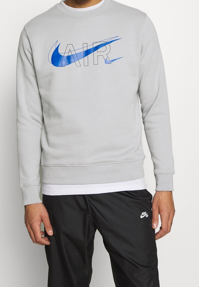 Grå sweatshirt med blå Nike "AIR"-logotyp. Rundad halsringning, långärmade, mjuk textur. Matchad med svarta byxor med en vit logotyp.
