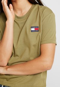 Olivově zelené bavlněné tričko s krátkým rukávem, které na levé straně hrudi zdobí vyšívané logo Tommy Jeans v modré, bílé a červené barvě.