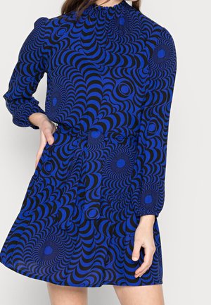 Femme portant une robe à manches longues avec un motif psychédélique bleu et noir, taille cintrée et col haut.