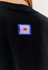 Czarna koszulka z kolorową naszywką przedstawiającą wzór oka na falistym fioletowym tle, obramowana niebieskim ściegiem.