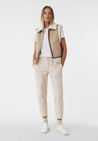 Beżowa bezrękawnik z kołnierzem z owczej wełny i bocznymi kieszeniami, w połączeniu z jasnobeżowymi spodniami i białymi sneakersami, na jednolitym tle.