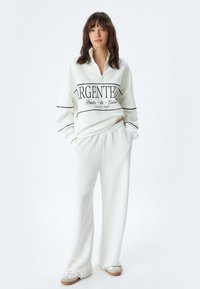 Sudadera blanca con cremallera y texto negro, acompañada de pantalones blancos de corte holgado. El modelo está de pie sobre un fondo neutro.