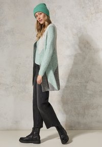 Lang, strikket cardigan i mintgrøn, lysegrå og mørkegrå, båret med en matchende minthat, sorte jeans og sorte snørestøvler.