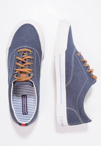 Blå duk sneakers med vita gummisulor, bruna snören och randig inredning. Har en strukturerad tyg och subtil logotyp.