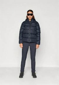 Kobieta w czarnej kurtce puchowej, ciemnych spodniach, butach turystycznych i dużych czarnych okularach przeciwsłonecznych, stoi na tle jasnego, jednolitego tła.