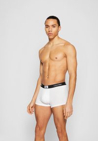 Balti medvilniniai bokseriai su juodu Calvin Klein juosmeniu ir logotipu, pasižymintys prigludusia konstrukcija ir lygiu paviršiumi.