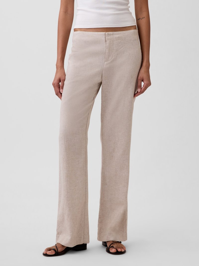 Femme portant un pantalon large en lin beige, un top blanc sans manches et des sandales plates noires à brides, debout devant un fond uni.