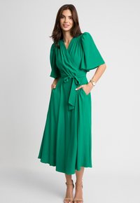 Zielona sukienka wrap z krótkimi bufiastymi rękawami, wiązana w pasie, z bocznymi kieszeniami. Wykonana z gładkiego, lekkiego materiału; długość do kostek, zwiewny krój.