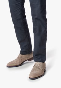 Chaussures en daim beige avec une texture tissée sur le bout, associées à un pantalon bleu marine foncé, offrant une coupe lisse et un design ajusté.