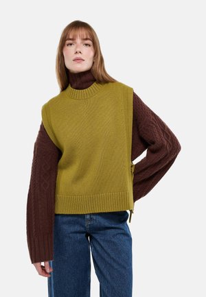 Femme aux cheveux bruns clairs et raides portant un pull à col roulé en maille torsadée marron sous un gilet sans manches en maille vert olive et un jean bleu.