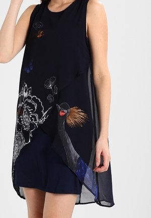Kvinde iført sort ærmeløs kjole med gennemsigtigt lag med fugle-, sommerfugle- og blomsterdesign, stående mod en lys baggrund.