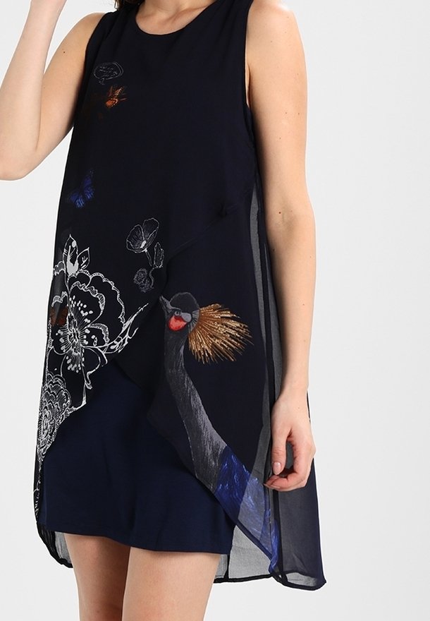 Femme portant une robe noire sans manches avec un voile transparent orné de motifs d'oiseaux, de papillons et de fleurs, debout devant un fond clair.