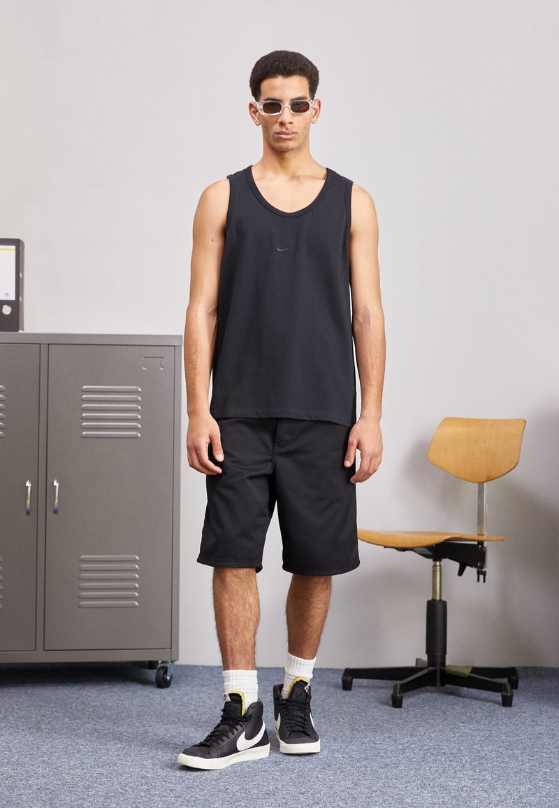 Camiseta sin mangas negra con cuello redondo, combinada con pantalones cortos negros. El calzado son zapatillas negras de caña alta con acentos blancos. Unas gafas de sol transparentes completan el look.