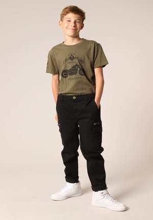 T-shirt vert olive avec graphique de moto et pantalon cargo noir avec poches, porté avec des baskets blanches.